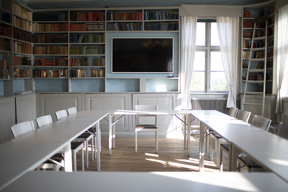 Konferensrum med långa, vita bord och stolar.  En stor skärm för presentationer och böcker syns i bakgrunden.